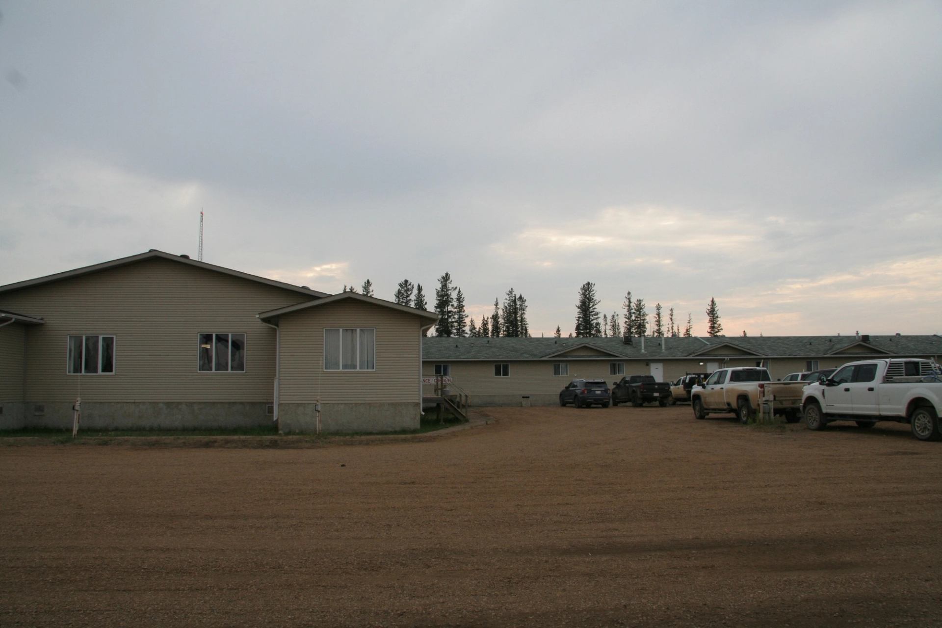 Single-storey beige buildings with parked pickup trucks on a wide gravel lot under cloudy sky.