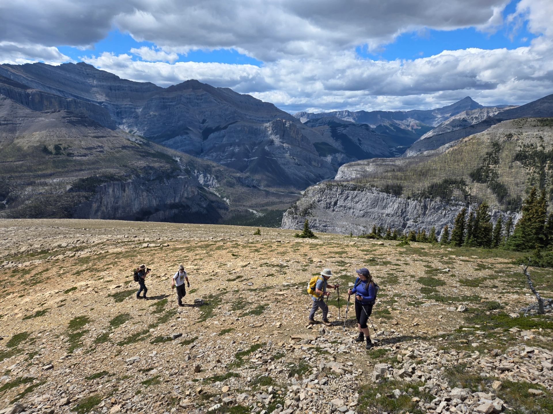 Hikers cross a rocky alpine slope with layered mountain ranges under scattered clouds.