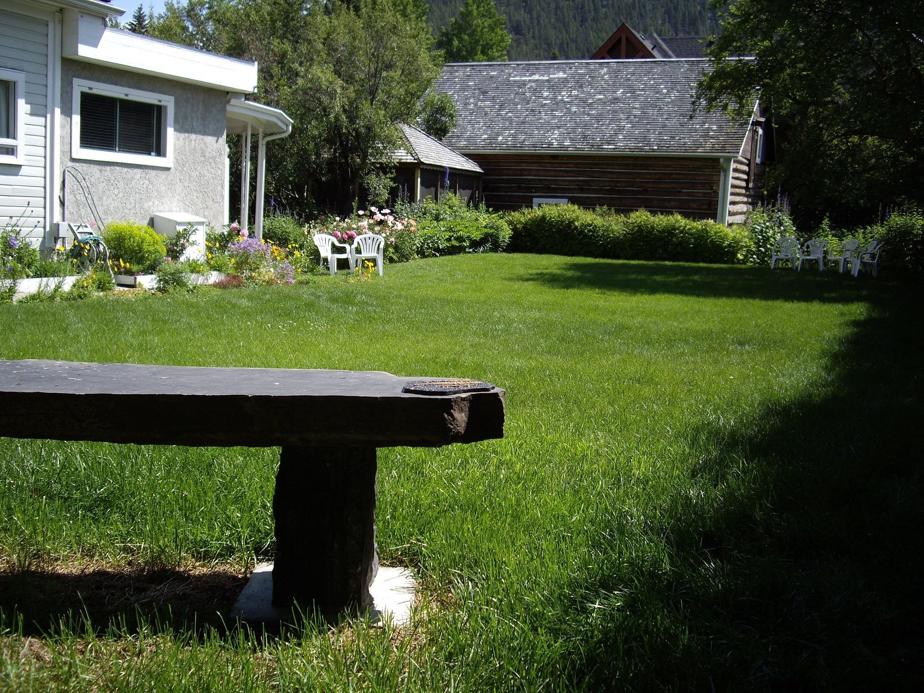 Garden lawn with white chairs, flowerbeds, and rustic buildings in a mountain landscape.