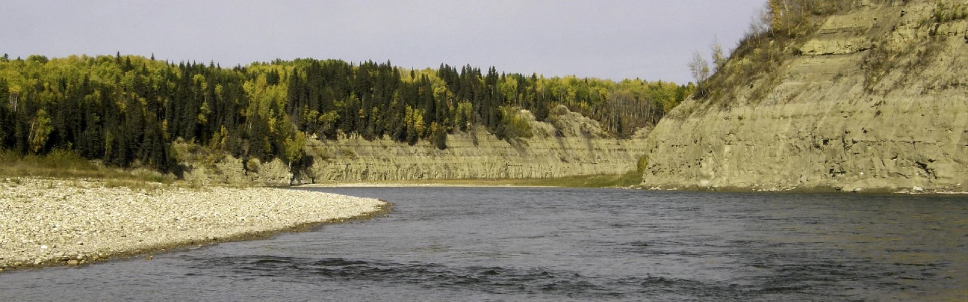 A river flows between a gravel bank, a forest with autumn colors, and a layered rock cliff.