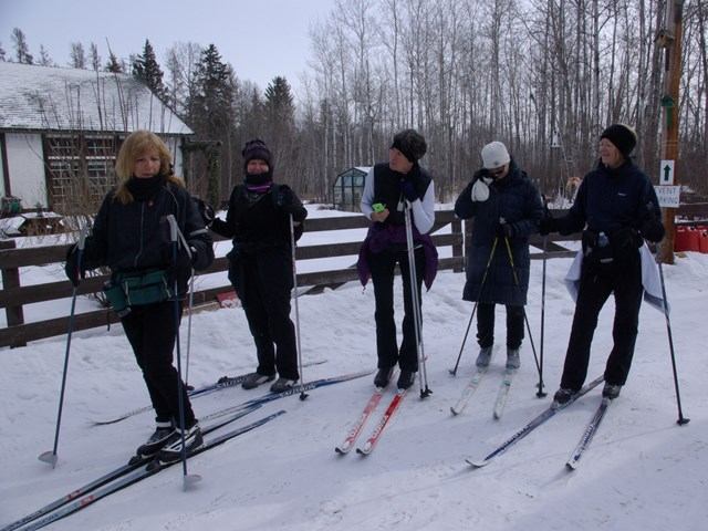 Group of skiers on snowy trail with gear, winter trees, and fence in the background.