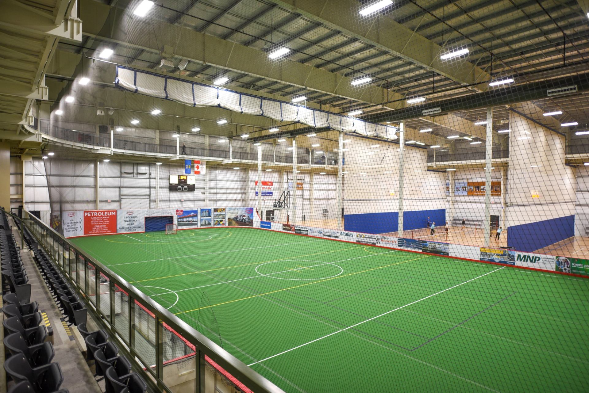 Indoor turf field with white markings, netting, and spectator bleachers.
