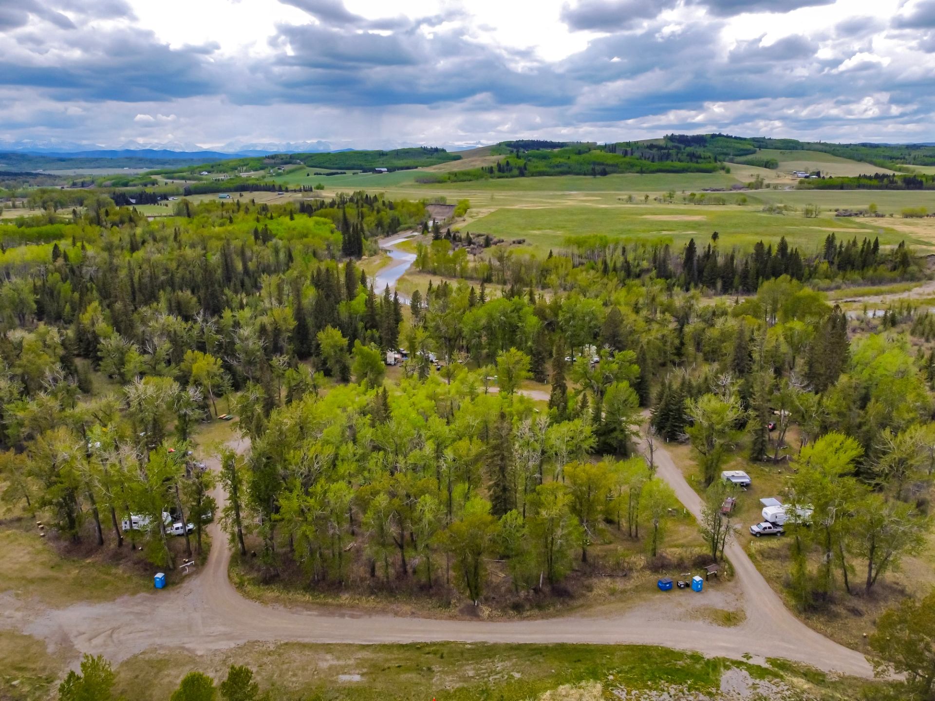 Aerial view of treed campground roads, river valley, and rolling hills under a cloudy sky near Millarville.
