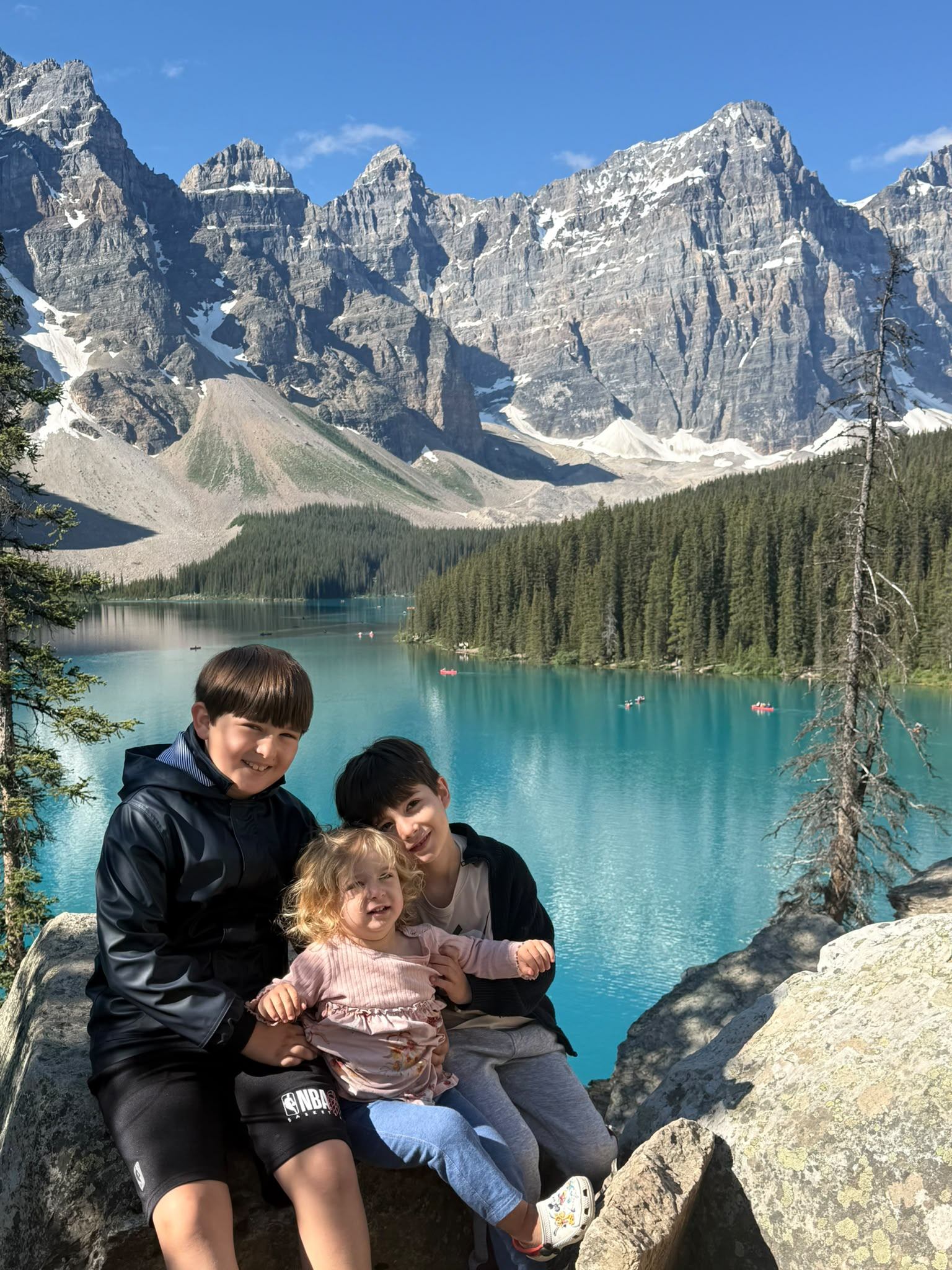 Three children sit on rocks overlooking a turquoise lake, forest and snow-capped mountains.