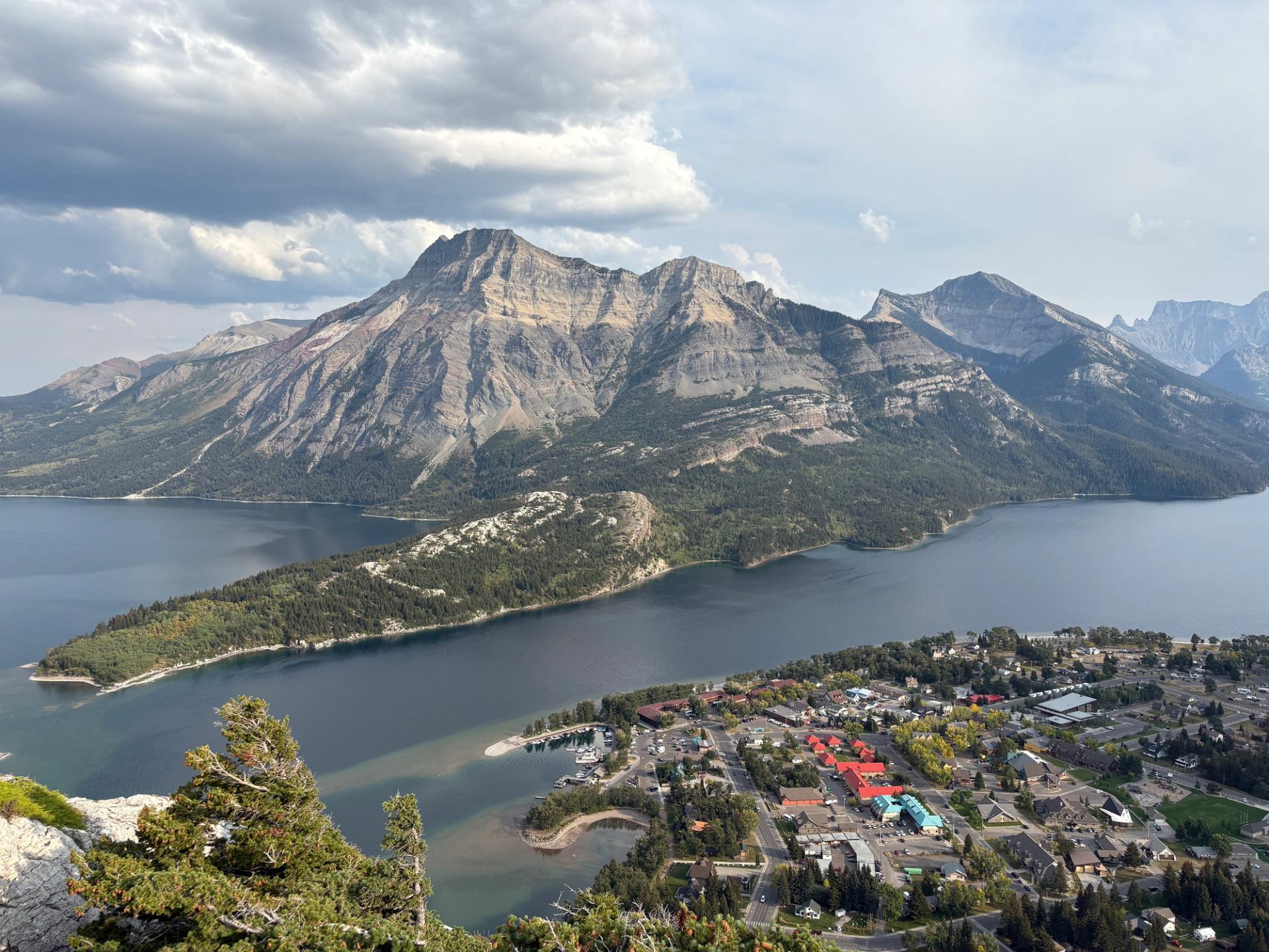 Mountain lake with forested slopes above a small town and marina under cloudy sky.
