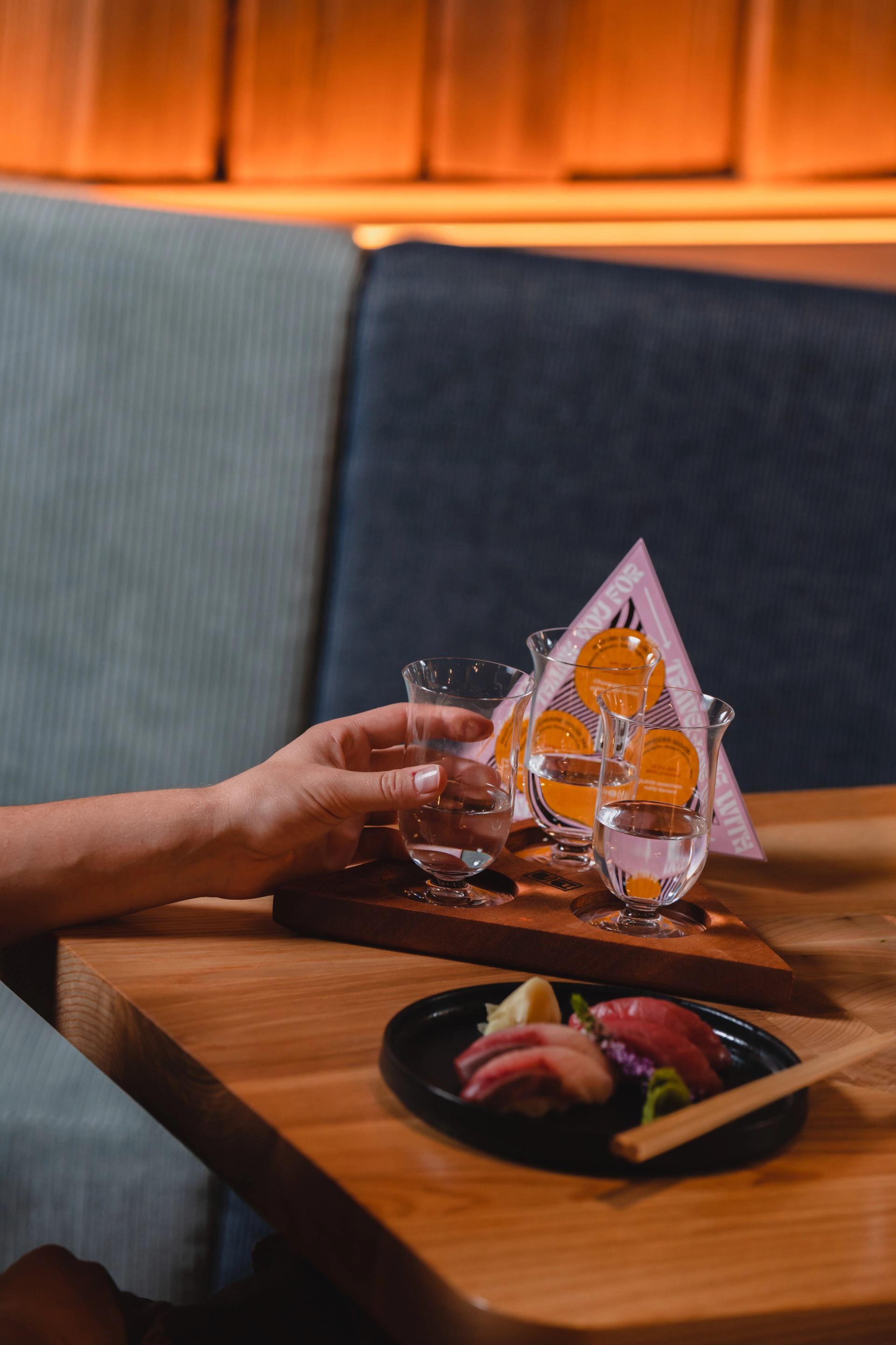 A hand holds a glass from a sake tasting flight next to a plate of sushi on a wooden table.