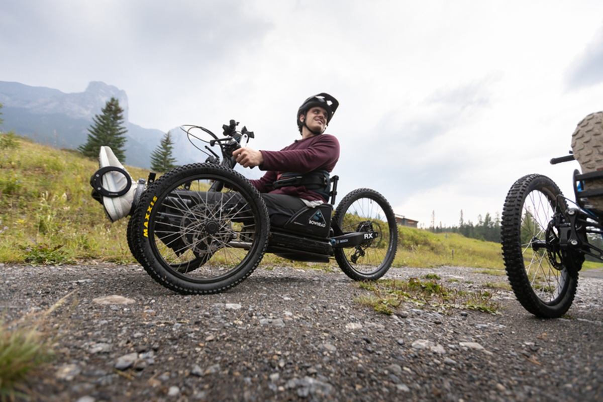 Person riding an off-road adaptive trike on a gravel trail in a mountain landscape.