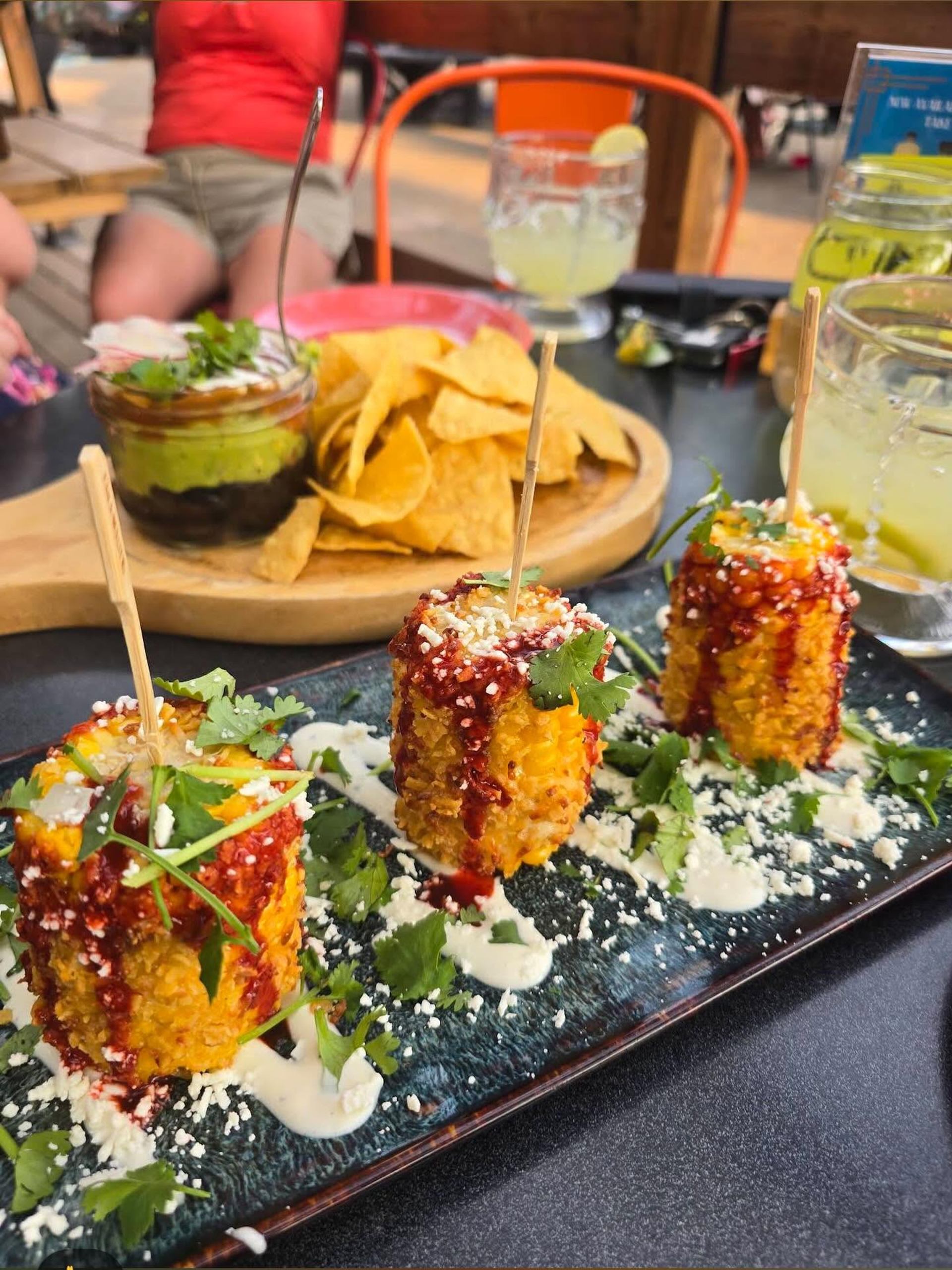 Plate of grilled corn skewers with sauce and cheese beside nachos and layered dip.