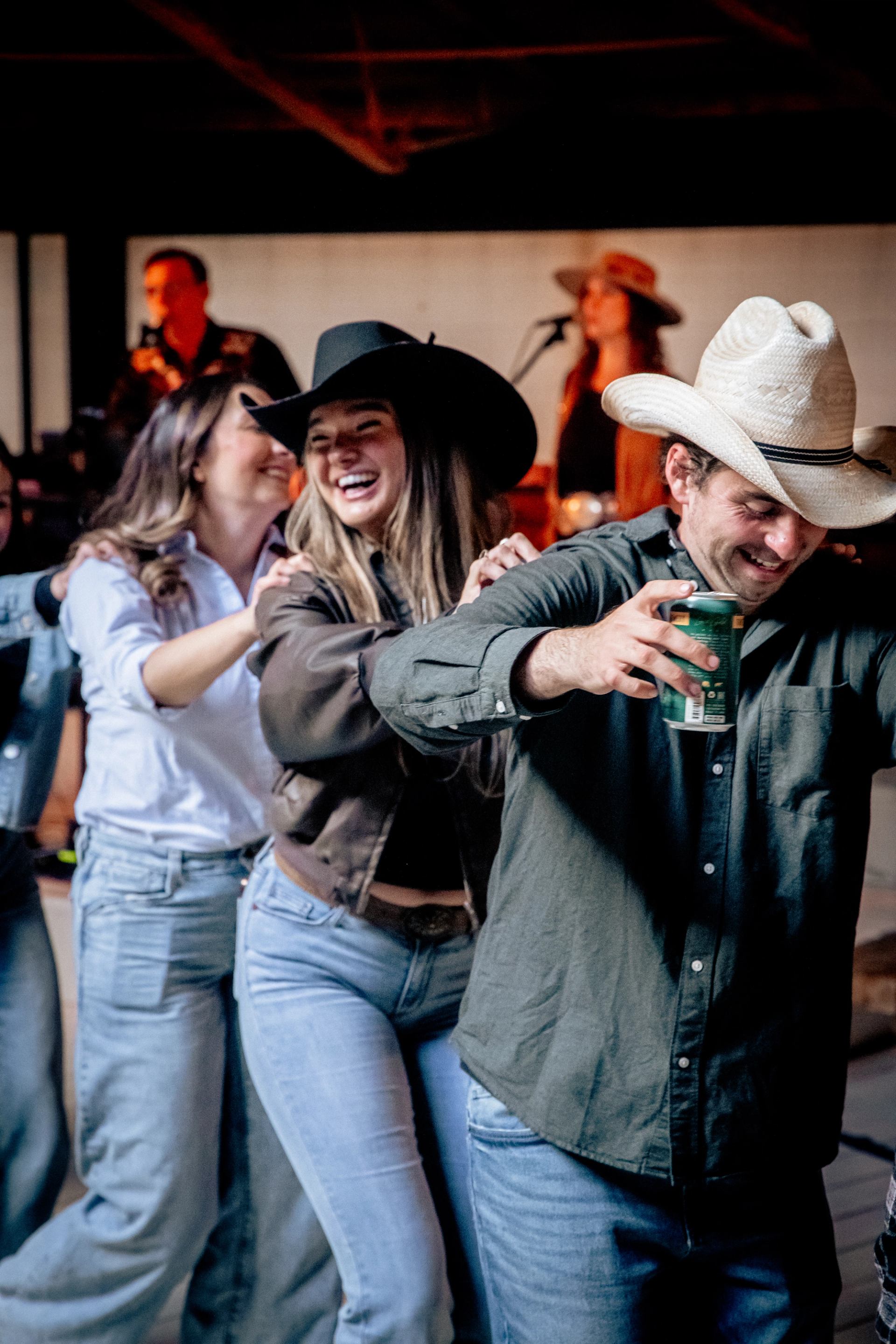 A joyful group in cowboy hats, arms linked, dancing and laughing with a band blurred in the background. 