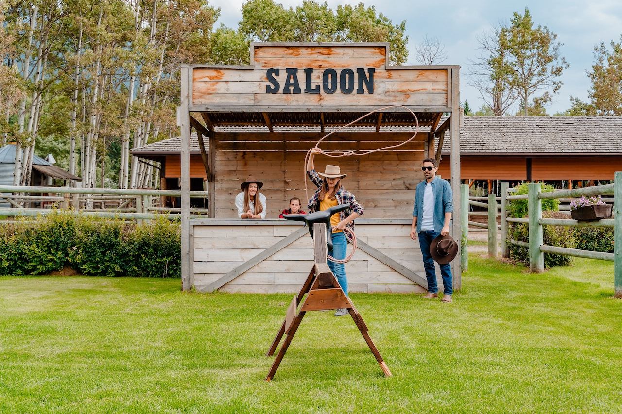 People practicing lasso skills in front of a wooden saloon backdrop on a grassy lawn.