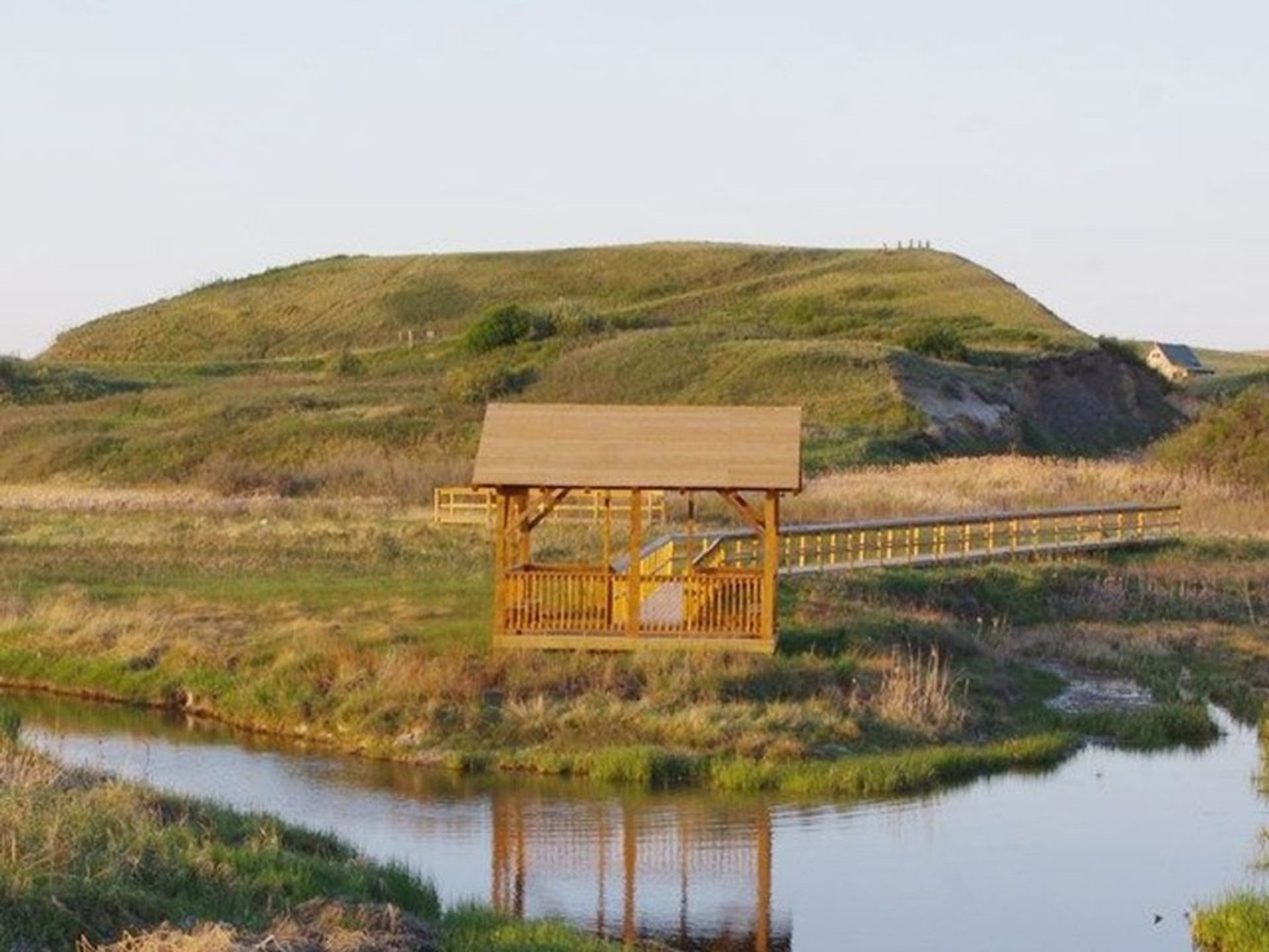 Wooden gazebo and boardwalk beside calm river, with grassy hills in background.