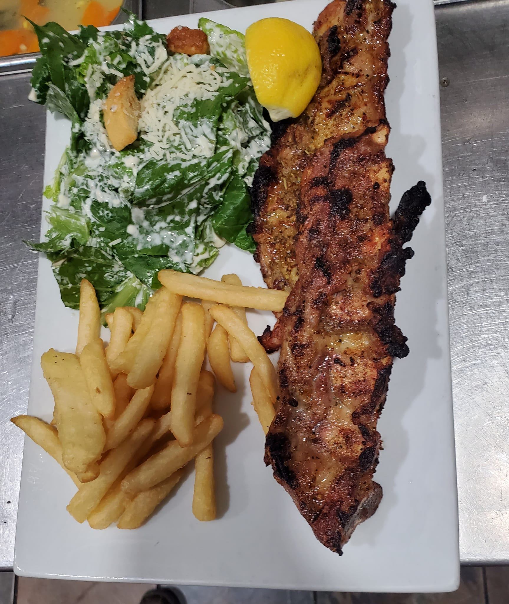 Grilled meat with French fries, Caesar salad and lemon wedge on white plate.