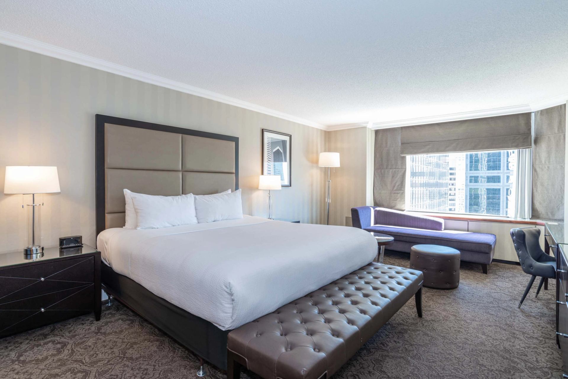 Hotel room with king bed, bench, purple sofa and city view window.