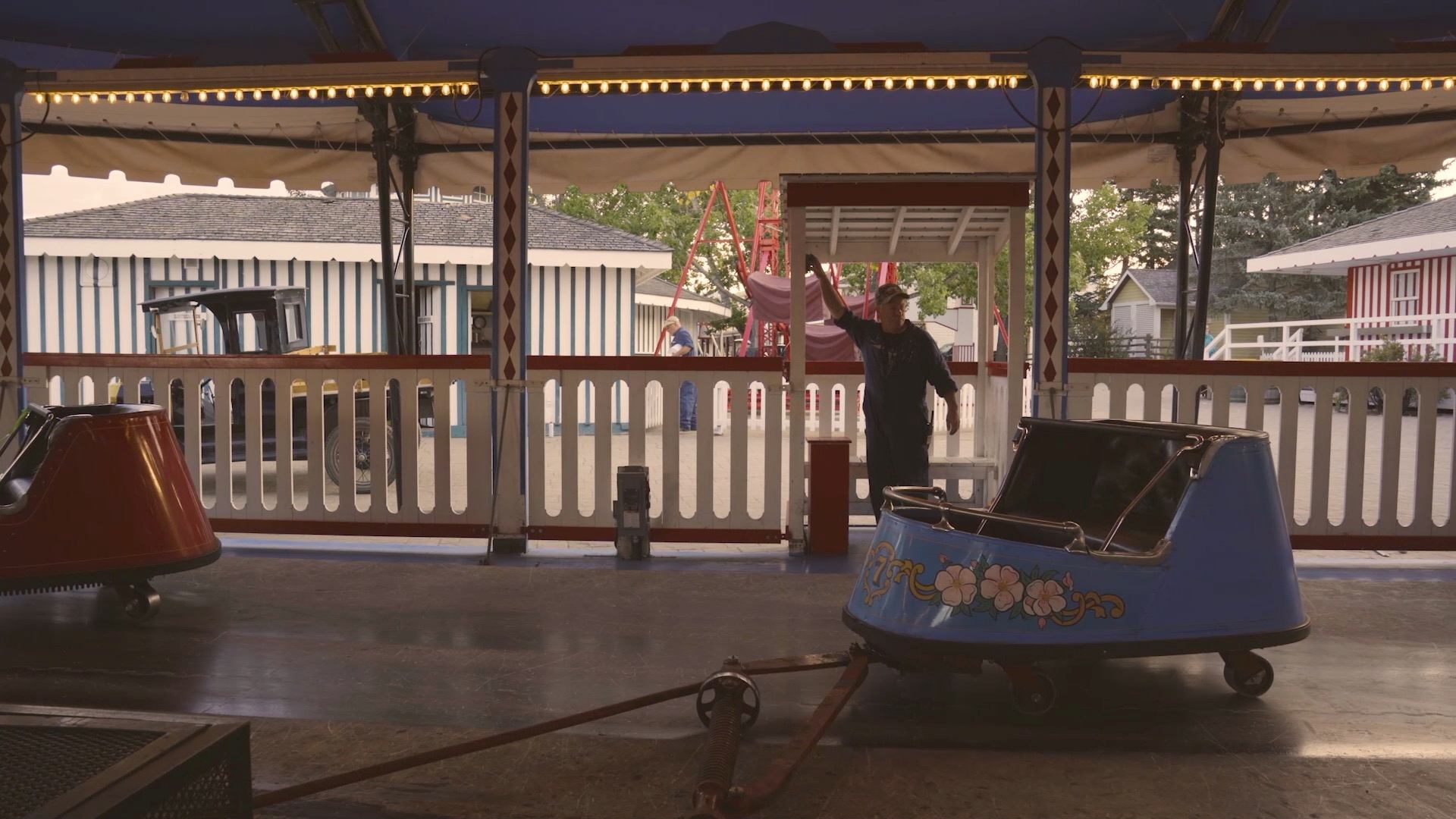 Amusement park worker stands beside empty blue ride car under covered pavilion.