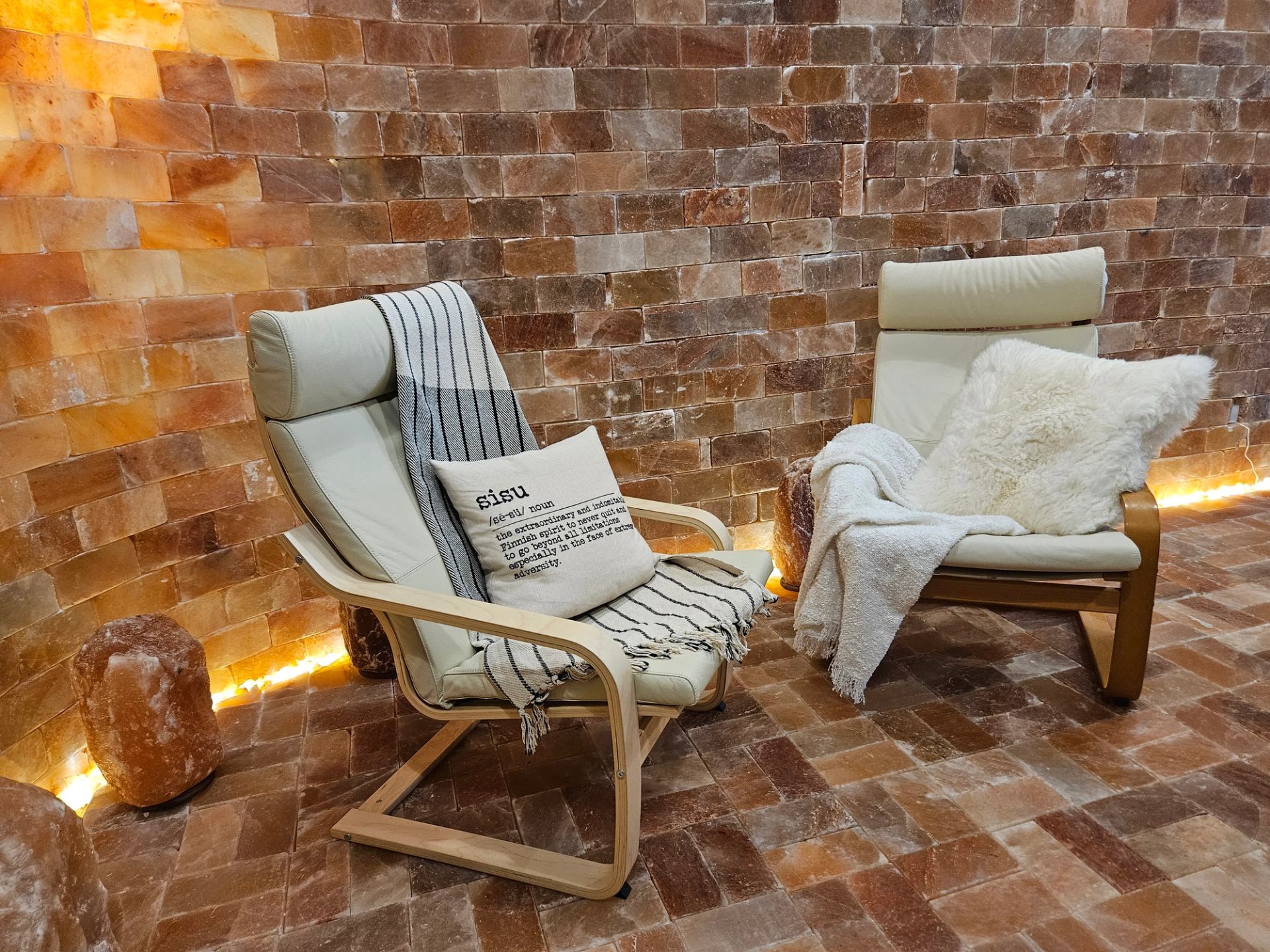 Two cozy cream lounge chairs equipped with plush blankets and pillows rest against a warm Himalayan salt brick wall. Soft under-lighting illuminates the salt floor, creating a serene relaxation space.