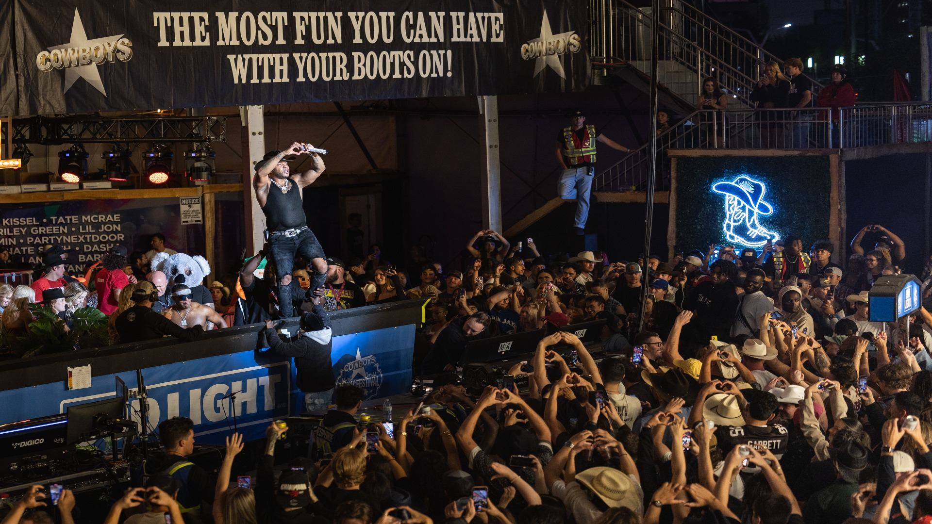 A male performer on an outdoor stage sings to a large crowd making heart gestures, with a "Cowboys" banner and a cowboy boot neon sign visible.