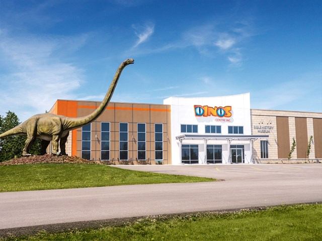 DINOS Centre Inc. building with dinosaur statue, parking lot and grass under blue sky.