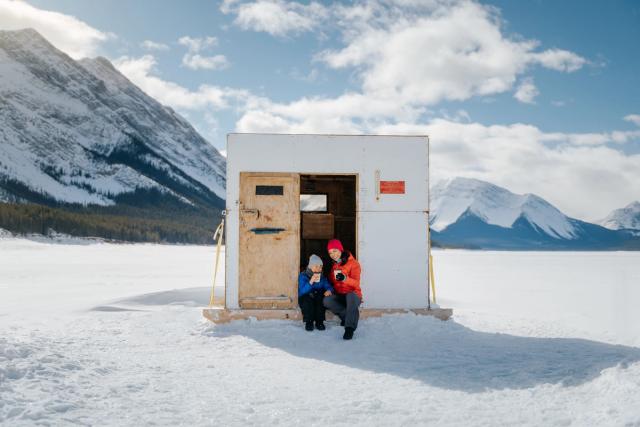 A mom and young child sit in the doorway of a ice fishing hut enjoying a hot beverage on a frozen lake near Canmore in Kananaskis Country