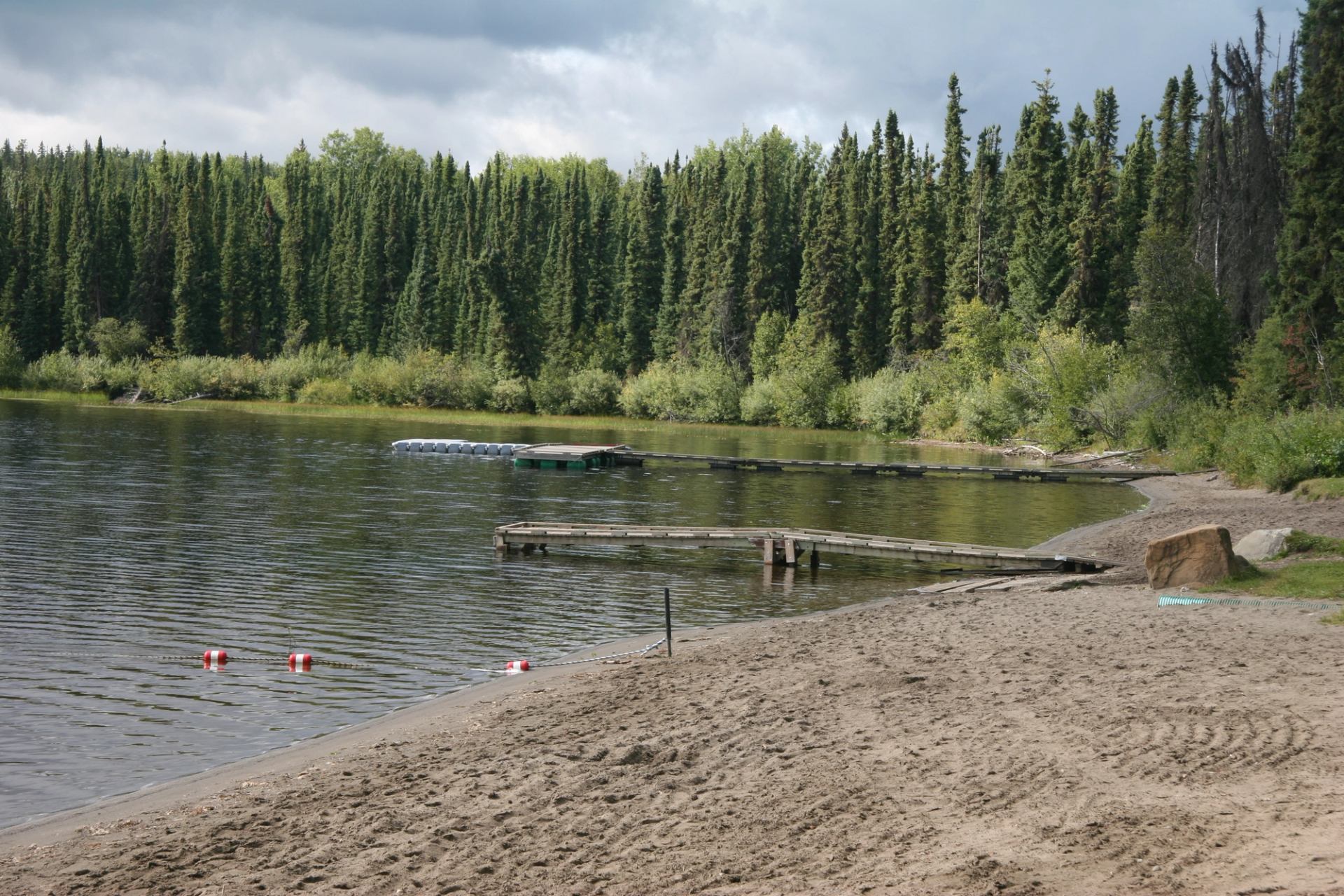 Sandy lakeshore with wooden docks, floating buoys and dense evergreen forest backdrop.