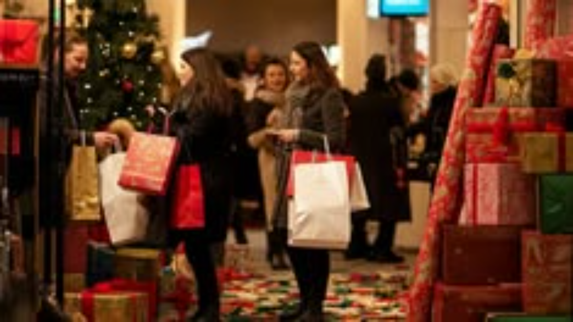Shoppers carry bags past a decorated tree and stacks of wrapped gifts.