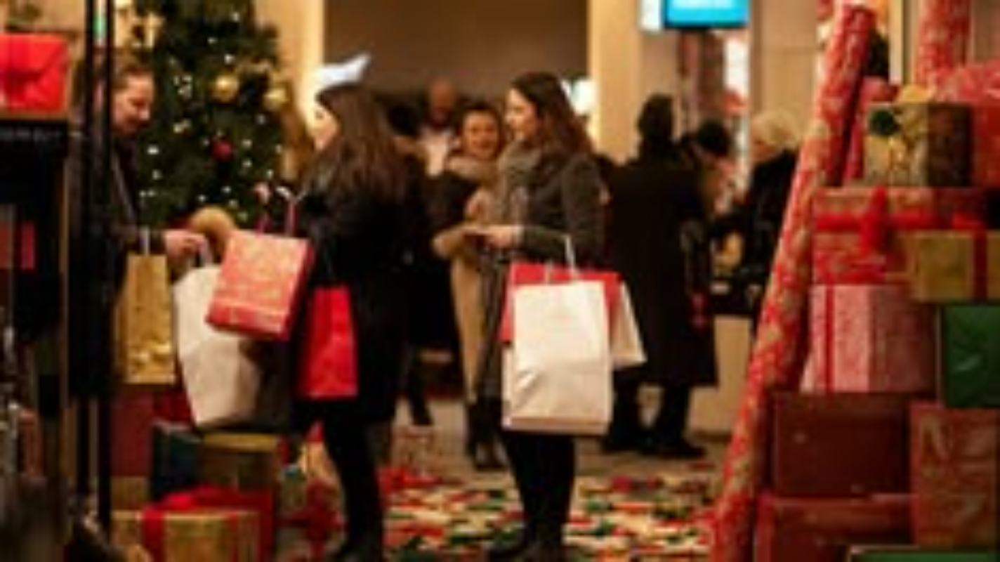 Shoppers carry bags past a decorated tree and stacks of wrapped gifts.