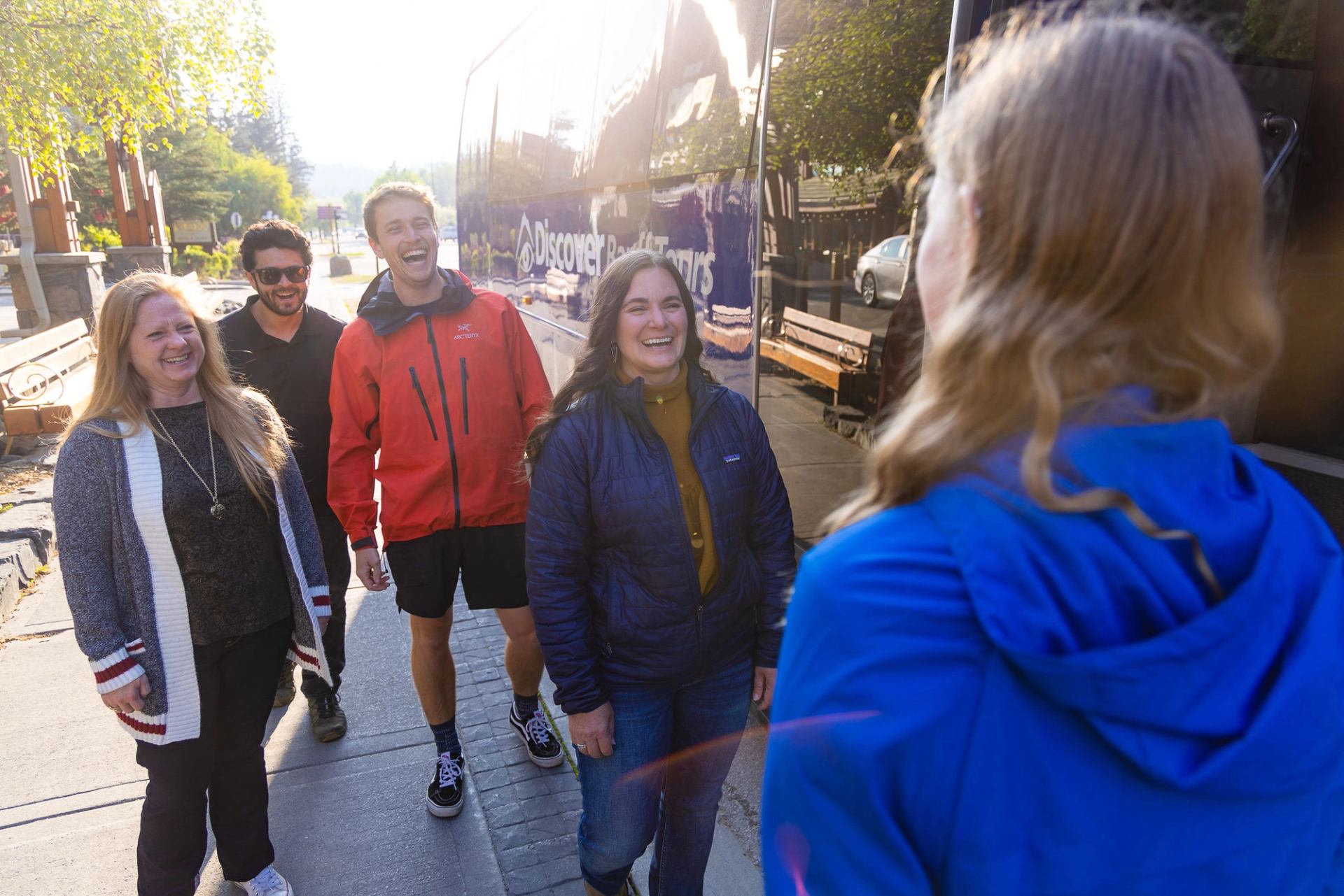 Group of people stand on sidewalk near tour bus, talking beside parked vehicles.