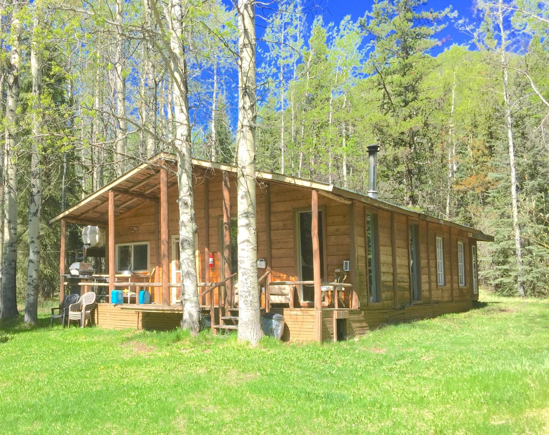 A log cabin surrounded by trees with a small deck, chairs, and a chimney on the roof.
