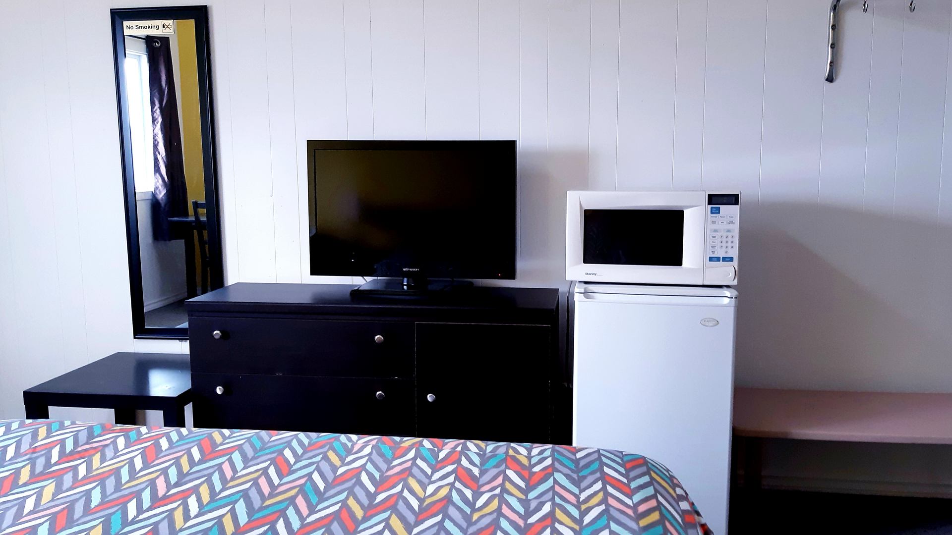 A guest room includes a television, mini fridge, microwave, mirror, and patterned bedding.