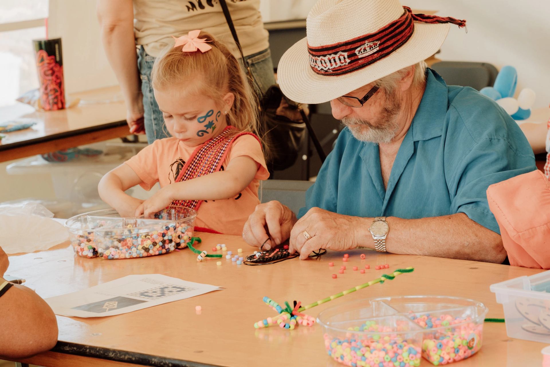 Child with face paint and older person make bead crafts at a table.