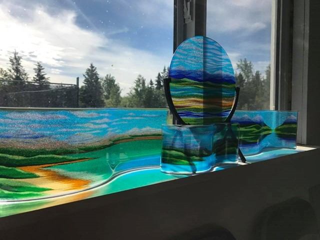 Coloured glass landscape pieces on a windowsill with trees and sky visible outside.