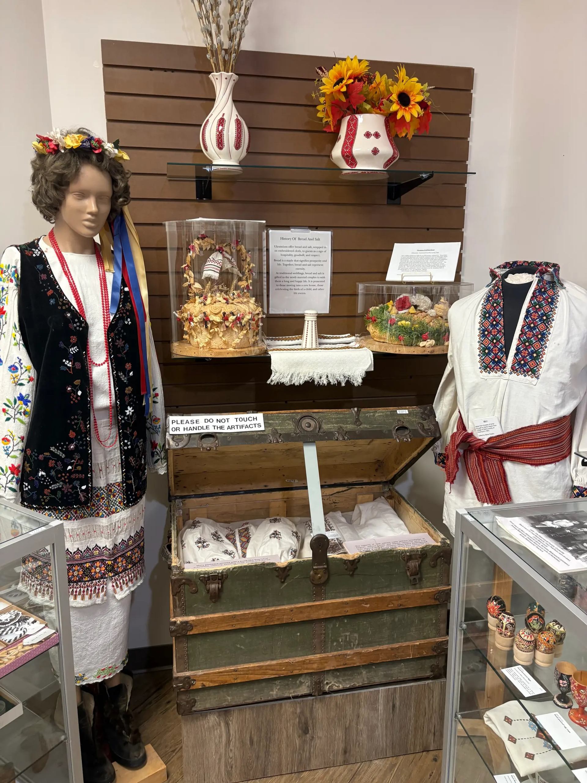Museum exhibit with mannequins in embroidered clothing, open trunk and display cases.