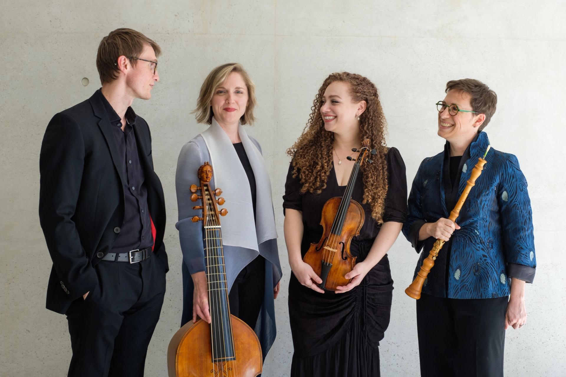 Four classical musicians stand against a light wall, holding string and woodwind instruments.