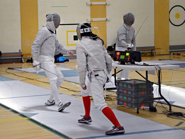 Fencers competing on a scoring strip with electronic scoring equipment in a gym.