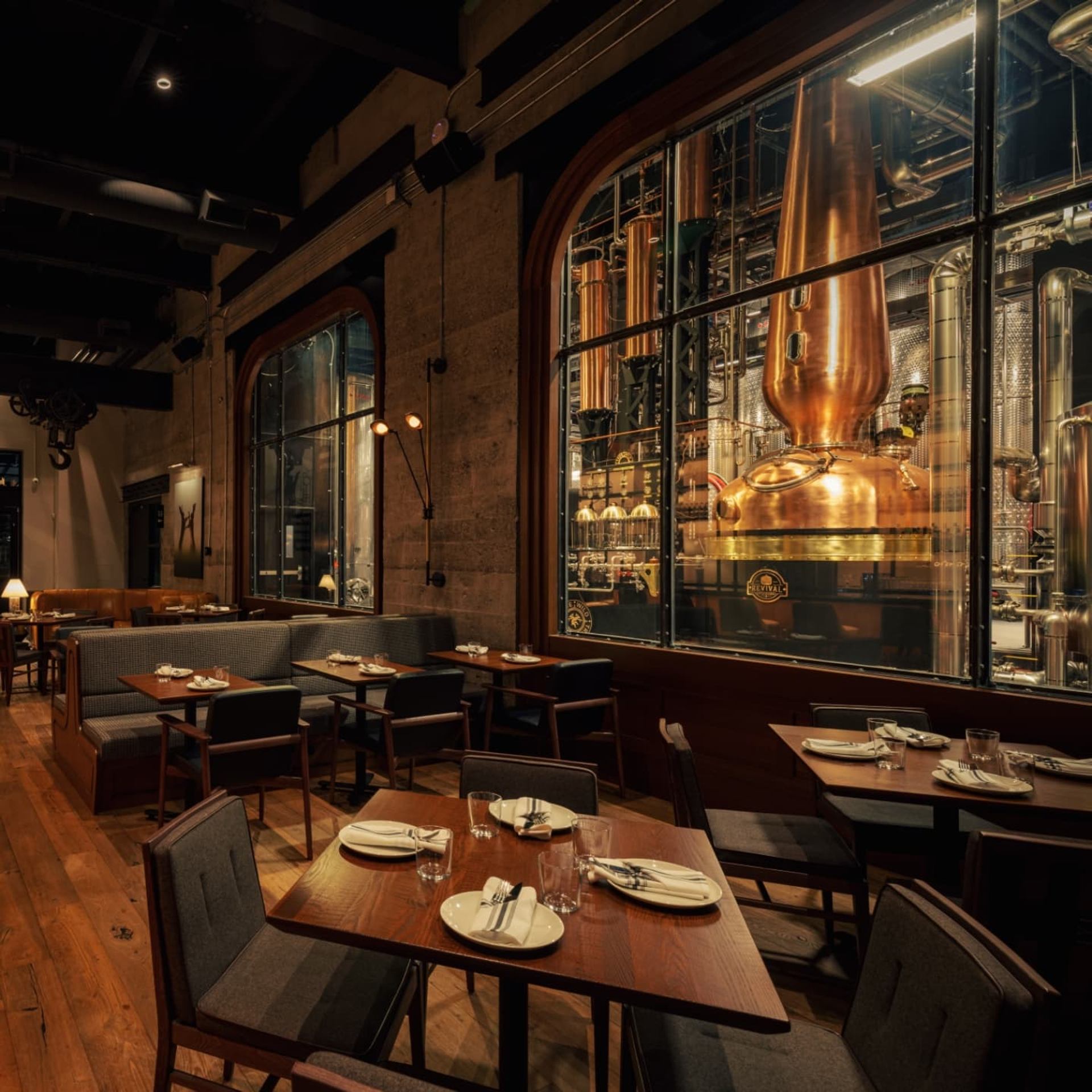 Dining room with wooden tables set for service, warm lighting, and large copper distilling equipment behind glass.