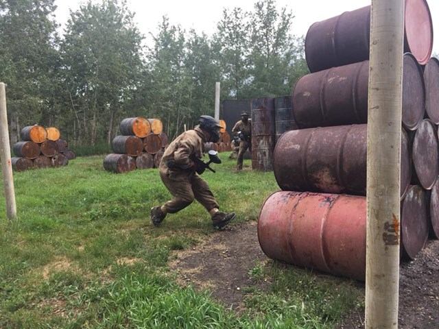 Paintball players run between stacked barrels on grassy field beside trees.
