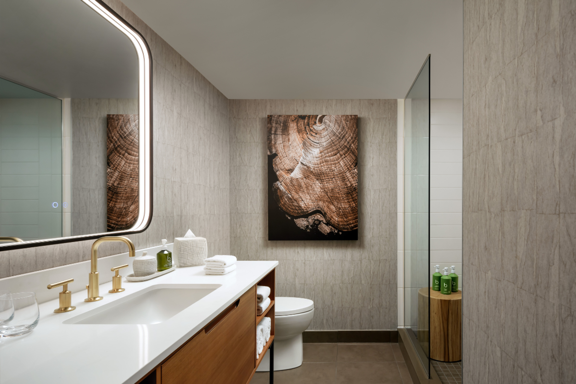 Modern bathroom with lit mirror, wood vanity, tree-ring artwork and glass shower.