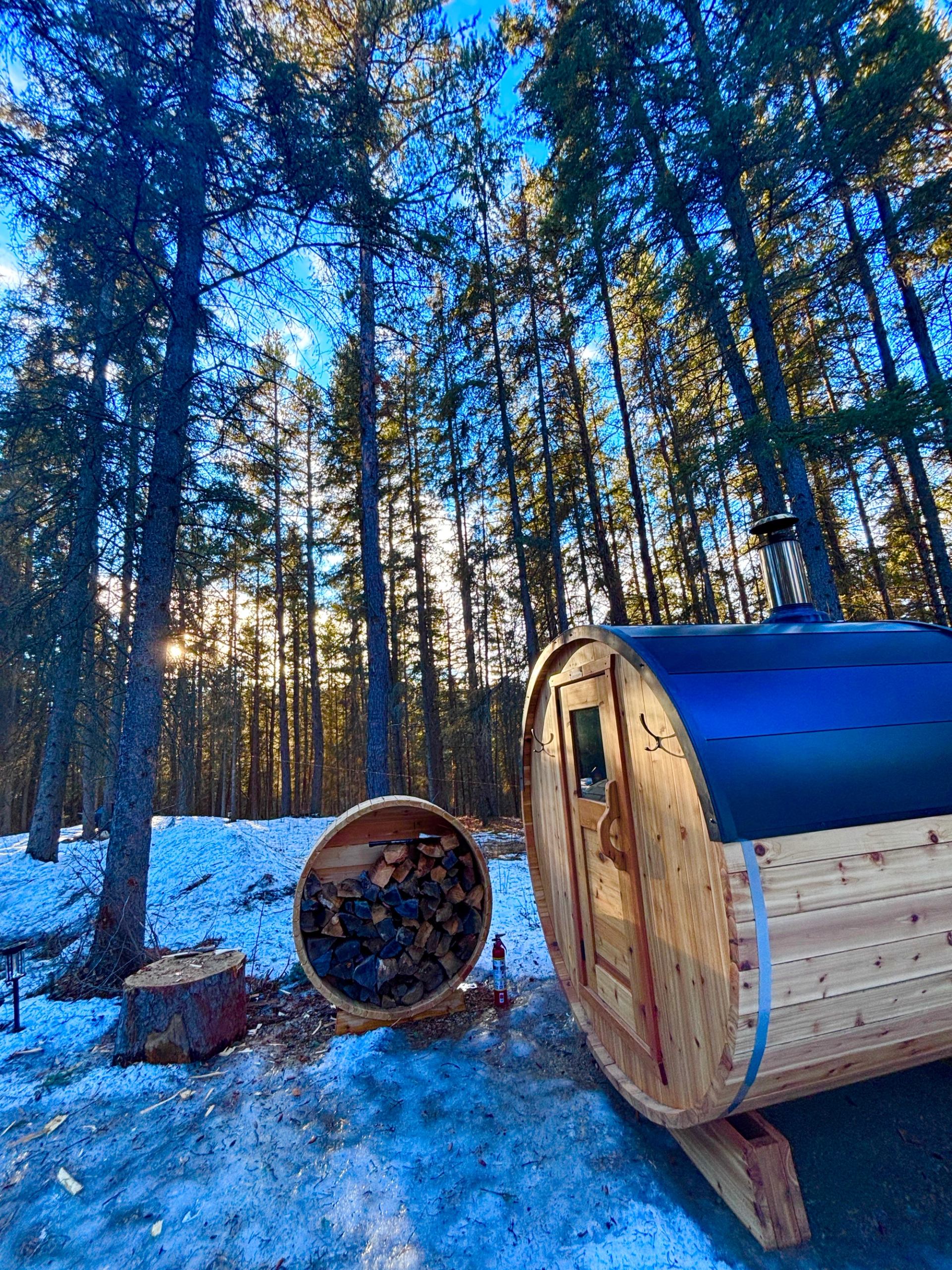 Wooden barrel sauna and round firewood rack in snowy forest clearing with tall trees.