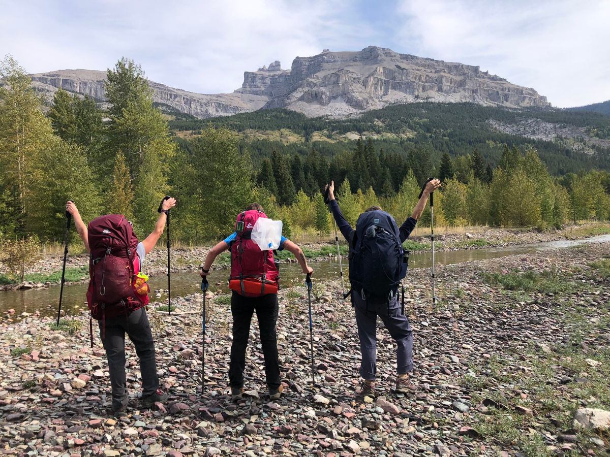 Three backpackers with trekking poles stand on rocky riverbank facing forested mountains.
