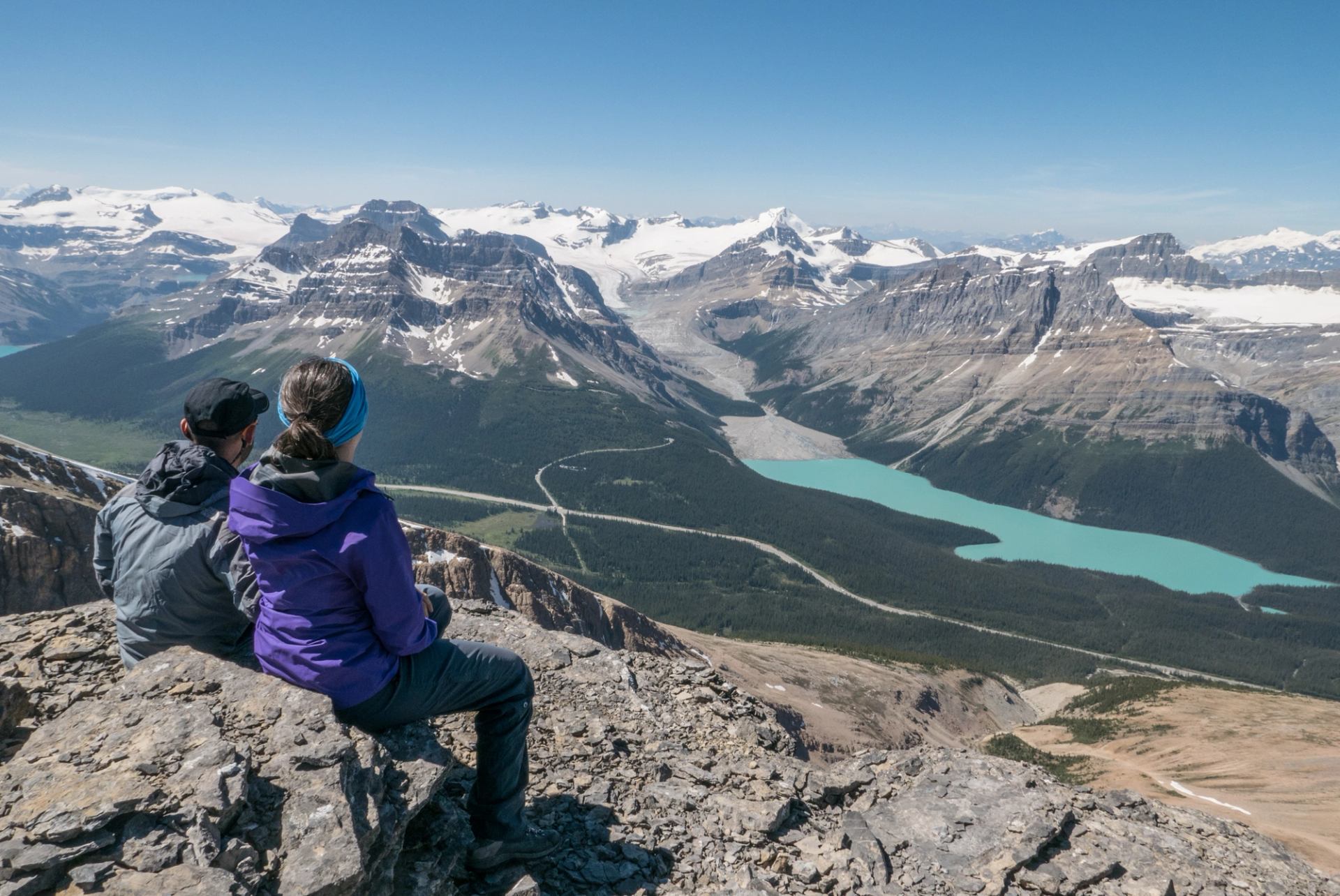 Two people sitting on rocky terrain overlooking snowy mountains and a turquoise lake.