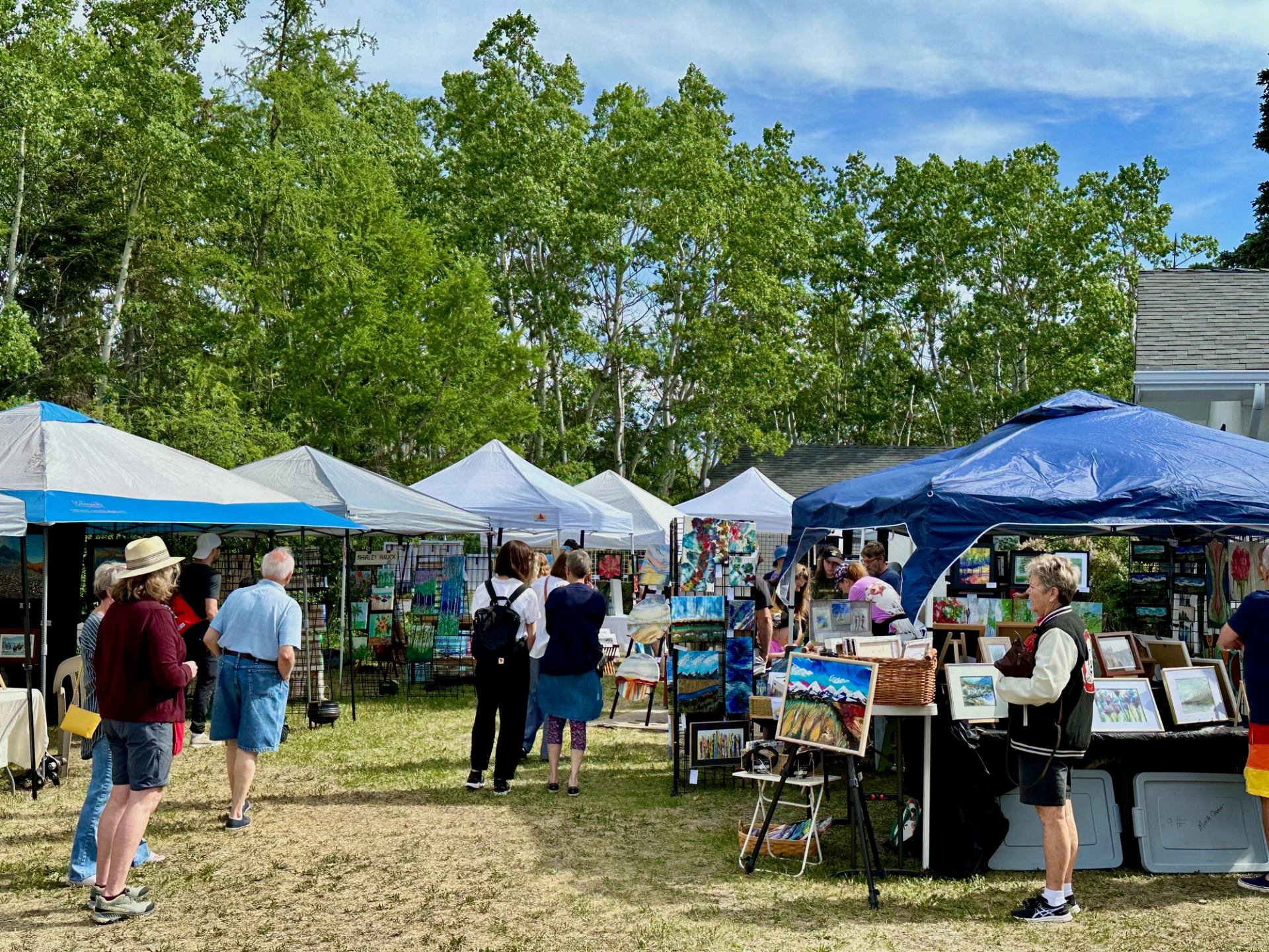 People browse art and crafts at an outdoor market with several tents on a sunny, grassy day.