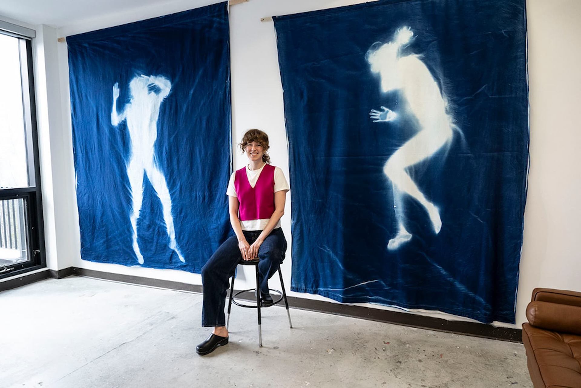 A woman sits on a stool between two large blue fabric hangings, each featuring a white silhouette of a human figure.
