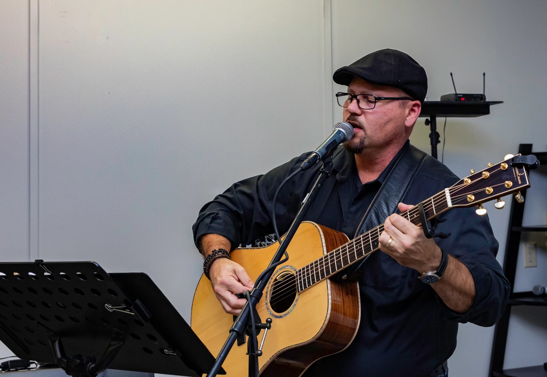 Musician with guitar and mic performing with sheet music and audio gear nearby.