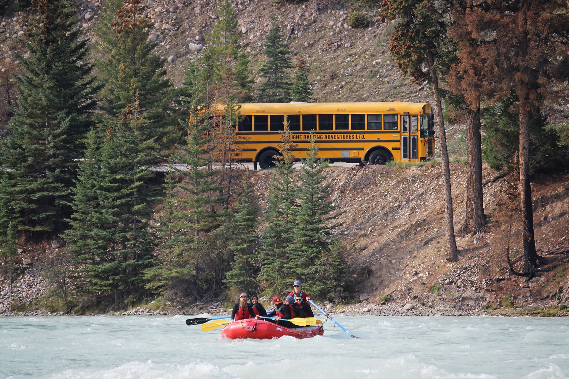 Red raft floating down a river with a yellow tour bus and forested hills in the background.