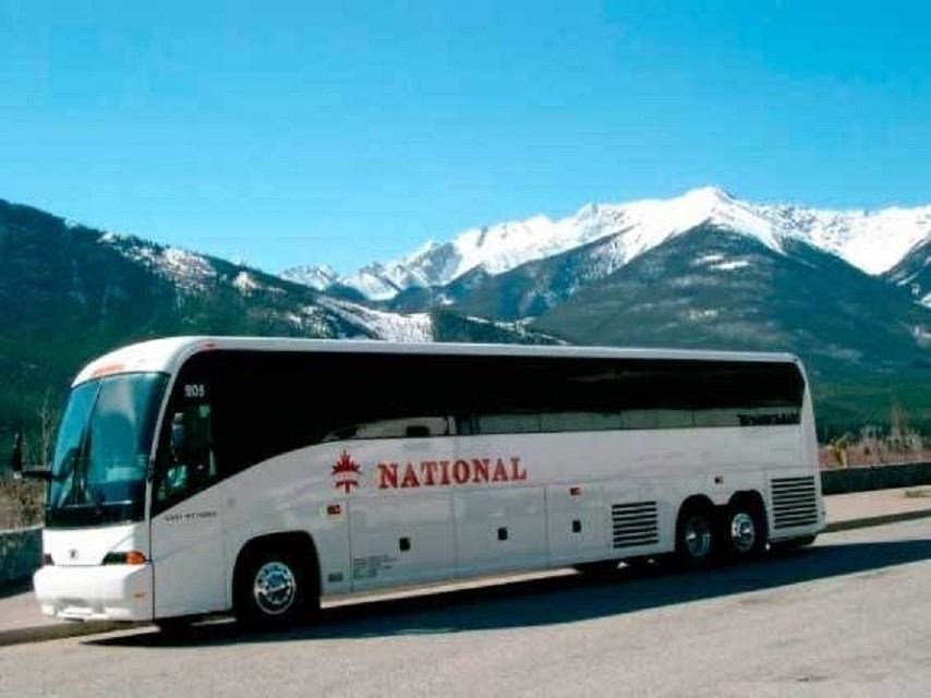 White coach bus with 'NATIONAL' logo parked near snow-capped mountains and blue sky.