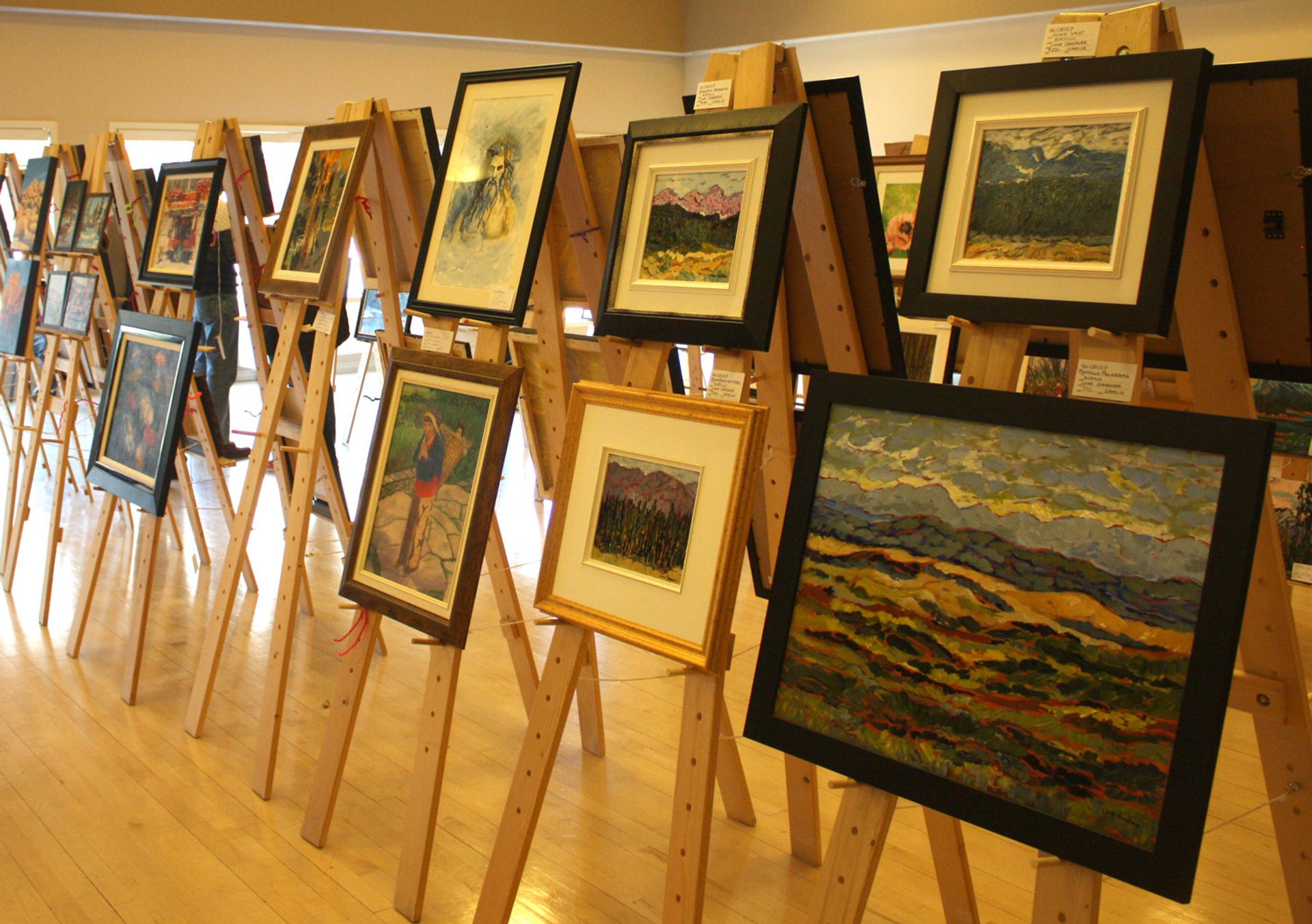Framed landscape and portrait paintings displayed on wooden easels in a bright gallery room.