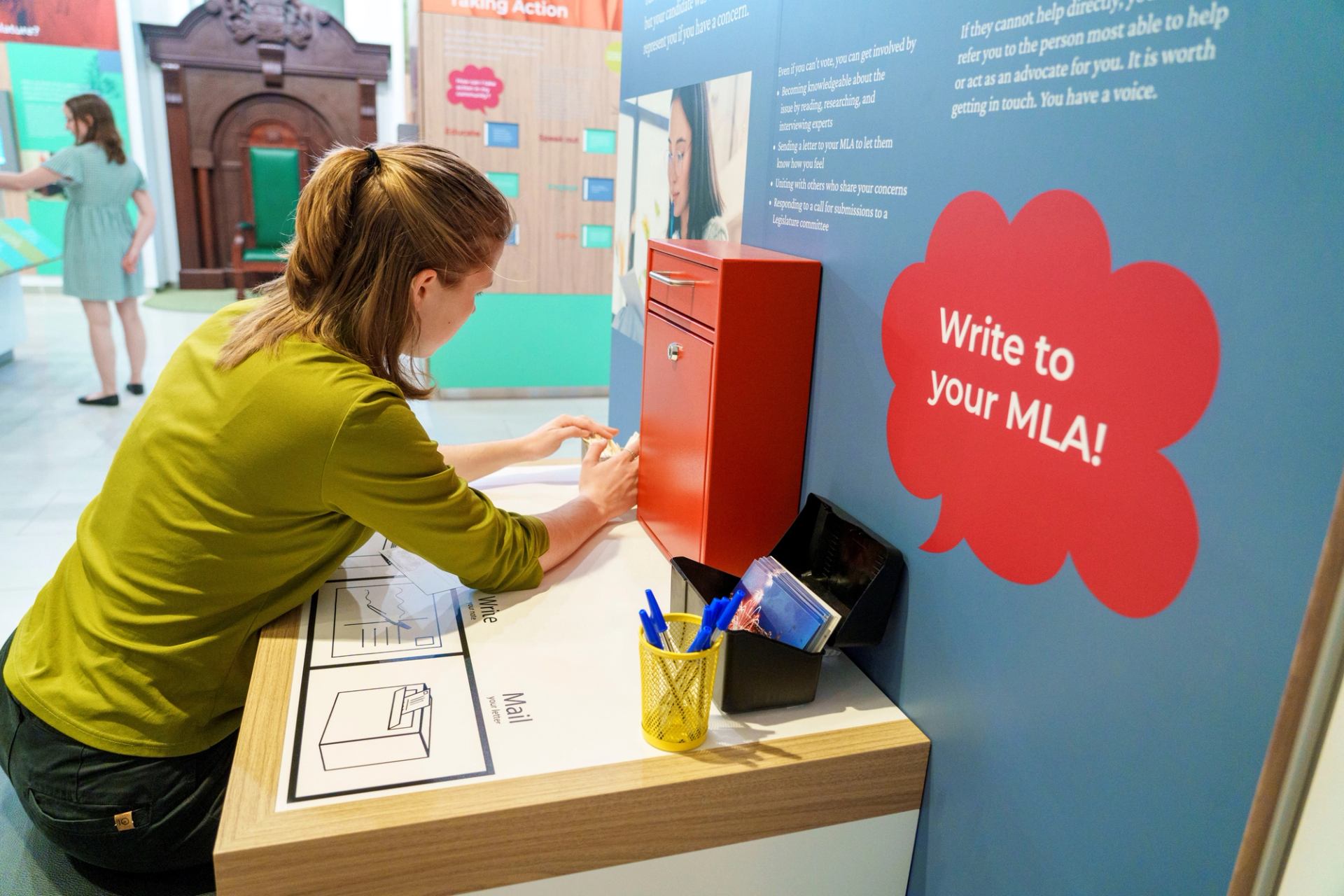 Person writing a letter at desk with red mailbox and advocacy sign.