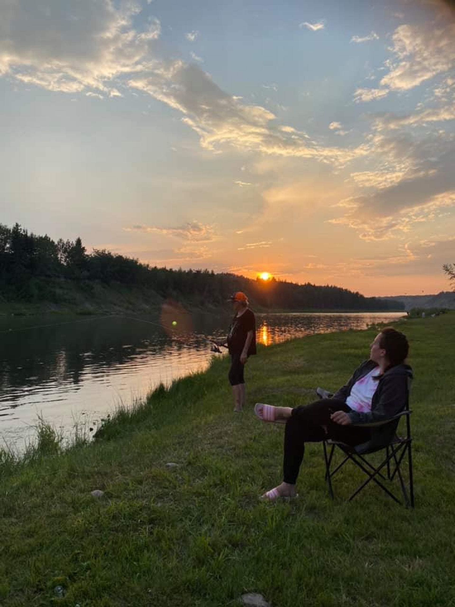 People relax beside a river at sunset with folding chairs along a grassy bank.