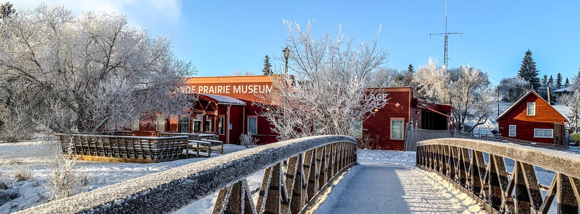 Snowy museum entrance with red building, frosty trees, and wooden bridge.