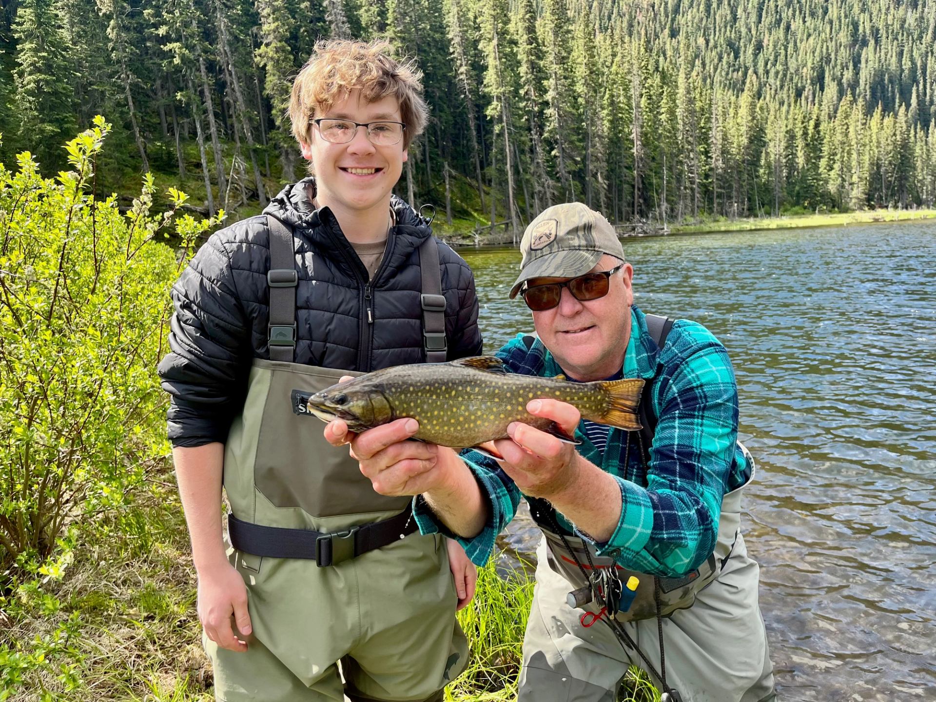 Two people in waders hold a speckled fish beside a forested mountain lake.