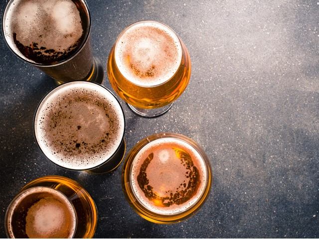 Five freshly poured beers with foam on a dark tabletop.